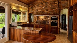 A kitchen with a brick wall, wooden cabinets, a built-in oven, and a large island with a round table. - Oak Hill Grand Country Escape with Sunset Views (Chirnside Park)