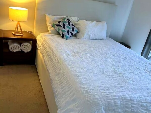A neatly made bed with a patterned pillow, a bedside lamp, and rolled towels on a nightstand.