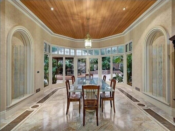 Un comedor amplio con techo de madera, ventanas grandes y una mesa de vidrio central rodeada de sillas.