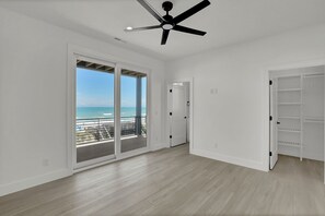 Una habitación amplia con ventilador de techo, puerta corrediza de vidrio que da a un balcón con vista al océano, y una puerta blanca con estante integrado.