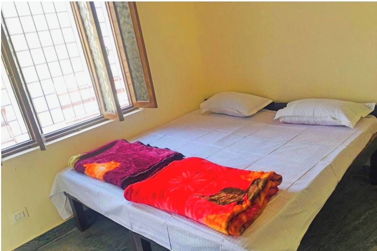 A single bed with a white mattress and two pillows, a window with metal bars, and a wall with a switch.