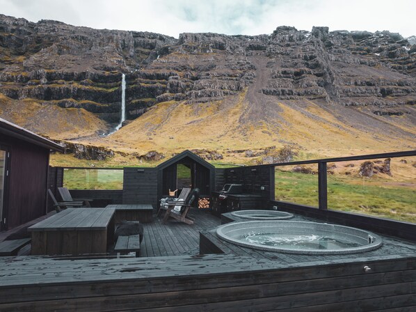 A wooden deck with a hot tub, lounge chairs, and a view of a waterfall and mountains.