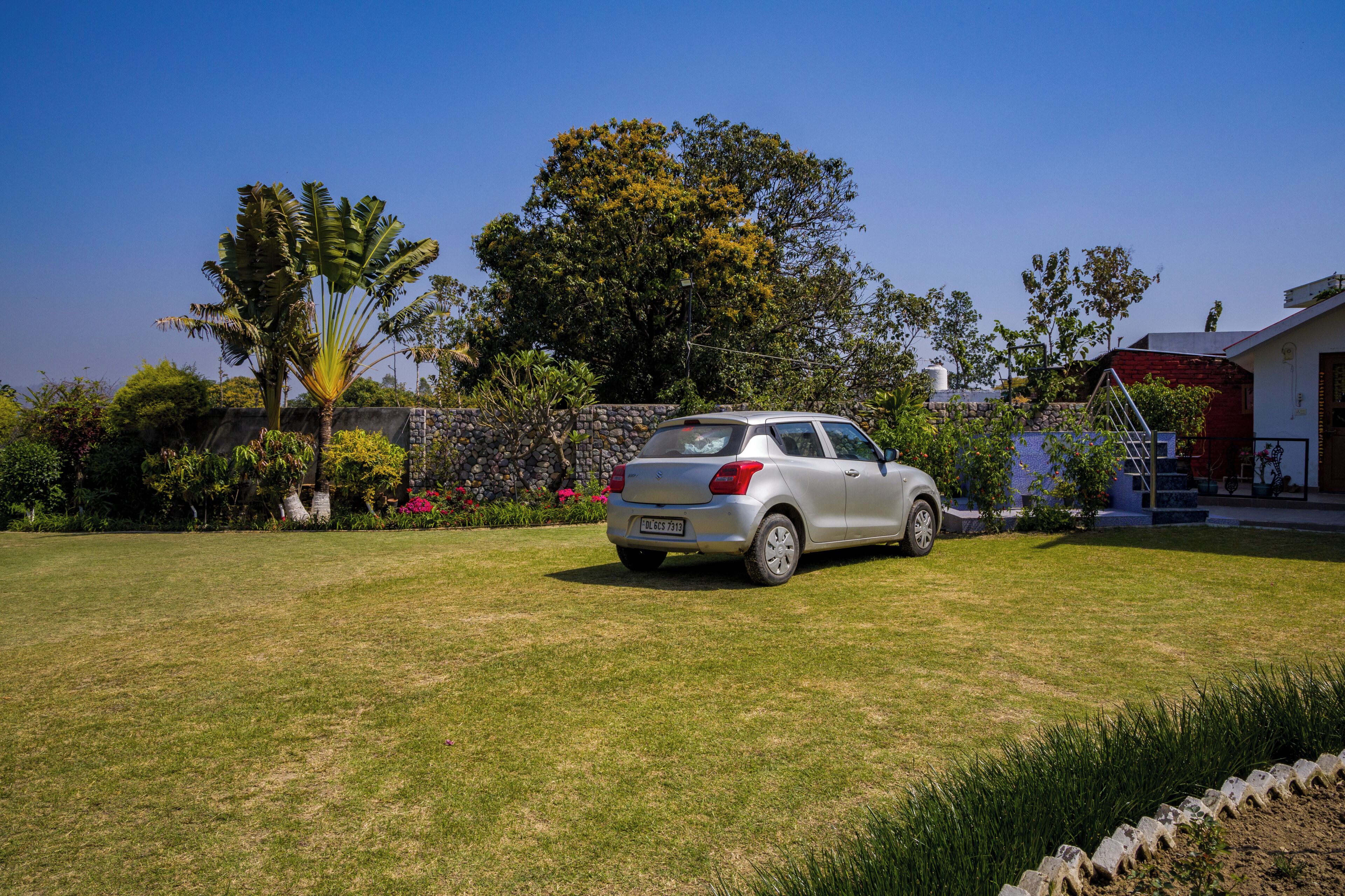 A car parked in a grassy area with a stone wall, trees, and a building in the background.