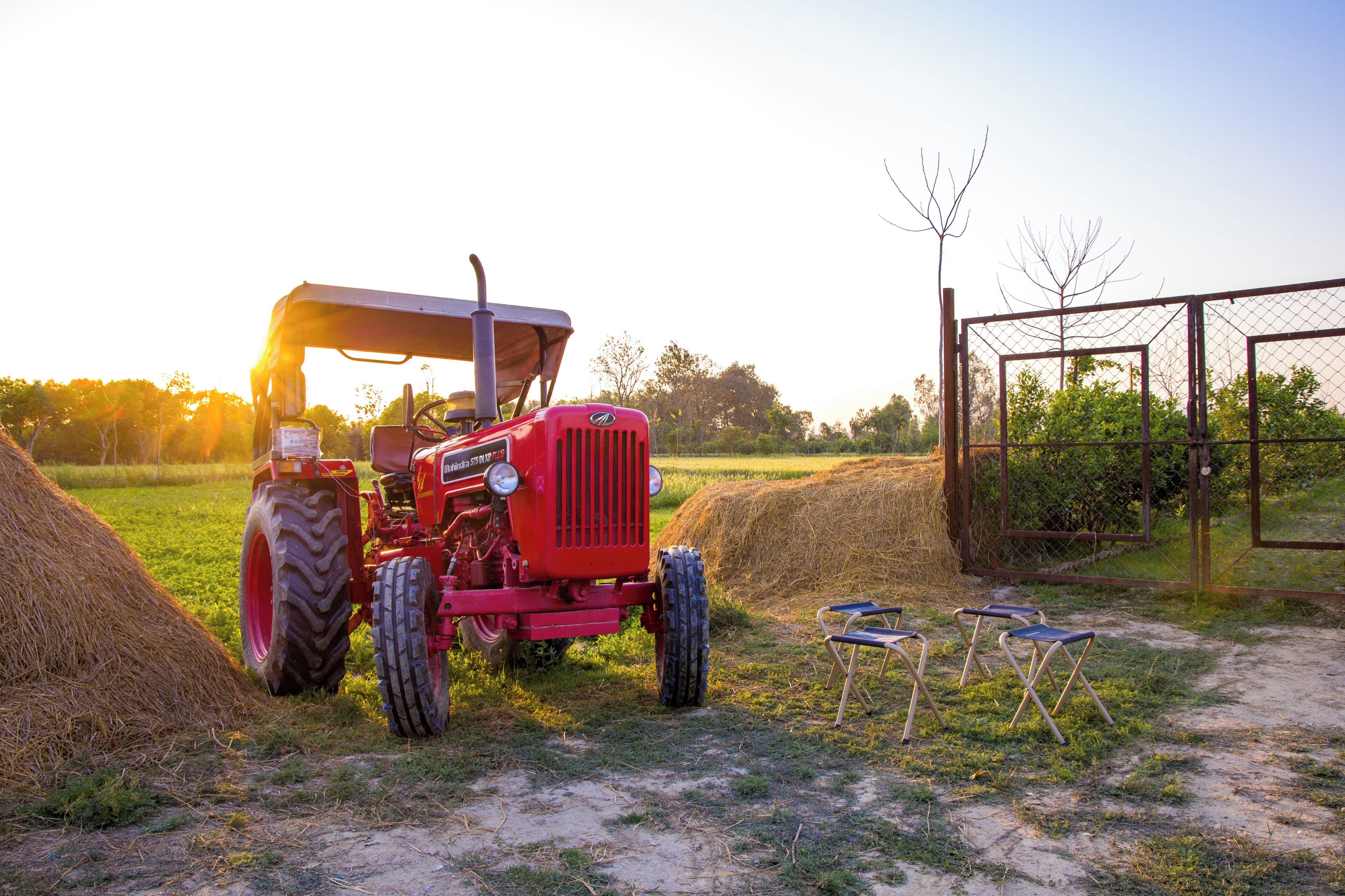 Un tracteur rouge avec une cabine, des balles de foin et deux chaises pliantes, le tout dans un cadre rural.