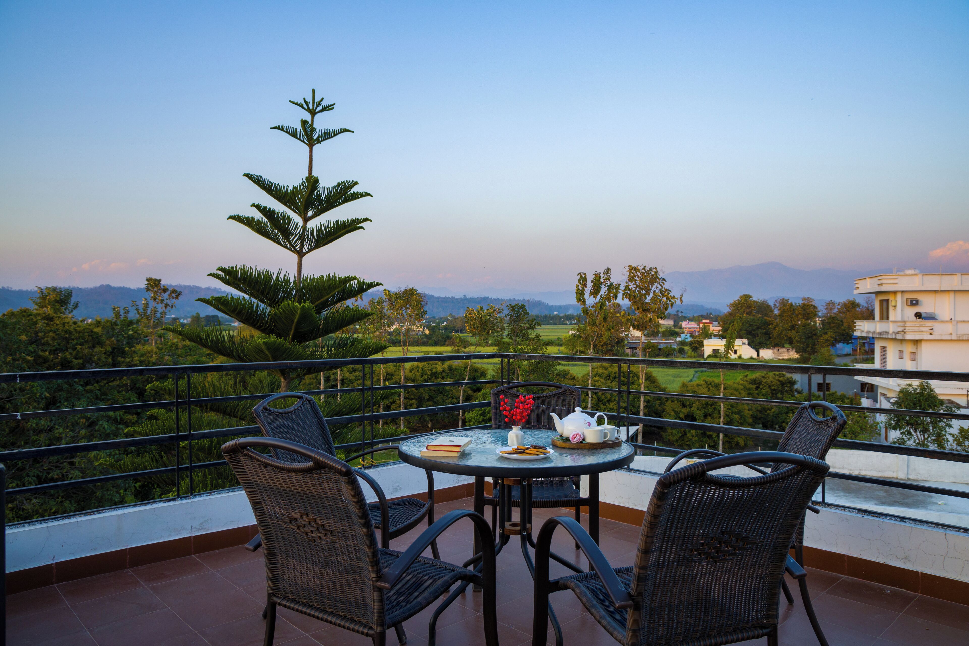Un balcon avec une table dressée pour deux personnes, des chaises, une plante en pot et une vue sur un paysage verdoyant avec des bâtiments visibles au loin.