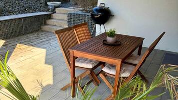 Een houten tafel en stoelen op een terras, met een barbecue, planten in potten en een stenen muur.