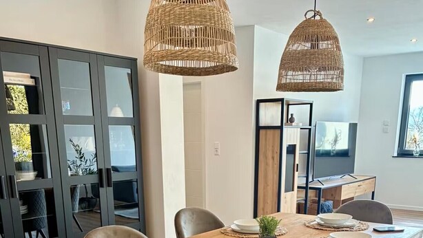 A dining area with a wooden table, six chairs, two hanging wicker lamps, and a glass partition with a view. - RaumWerk Apartment • family-friendly - Terrace (Wülfershausen an der Saale)