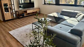 A modern living room with a grey sofa, a wooden entertainment unit, and a large window with a view of a house and trees. - RaumWerk Apartment • family-friendly - Terrace (Wülfershausen an der Saale)