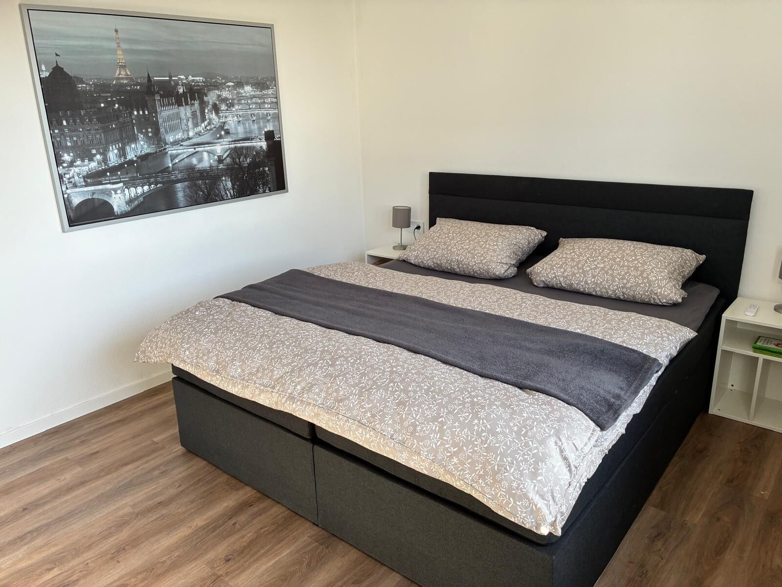 Une chambre à coucher avec un lit, une table de chevet, une lampe et un tableau représentant une vue sur la ville.