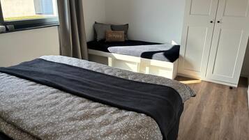 A bedroom with a bed, a small white desk, a wardrobe, and a window with curtains.