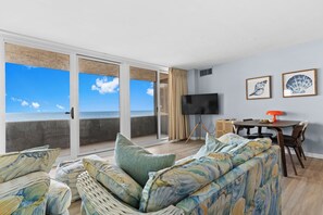 A room with a large sliding glass door leading to a balcony with a view of the sea, a flat-screen TV, a dining table with chairs, and a sofa with patterned cushions. - Antigua Oceanfront Retreat | 2BR + Heated Pool (Ocean City)