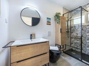 A modern bathroom with a round mirror, a white sink, wooden cabinets, a glass shower enclosure, and a plant. - Casa Sardinha - in the heart of Portimão (Portimão)