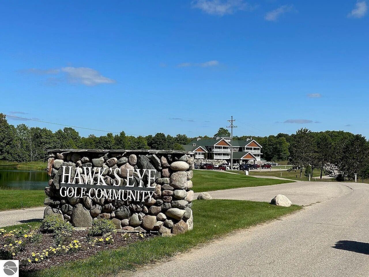 Un letrero de piedra con el texto 'HAWK'S EYE GOLF COMMUNITY', un camino de grava, un edificio con techo verde y una masa de agua al fondo.