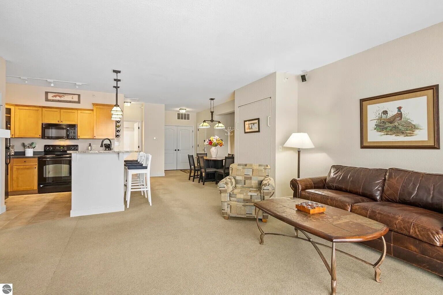 A living room with a brown leather sofa, a coffee table, and a dining area with a patterned armchair.