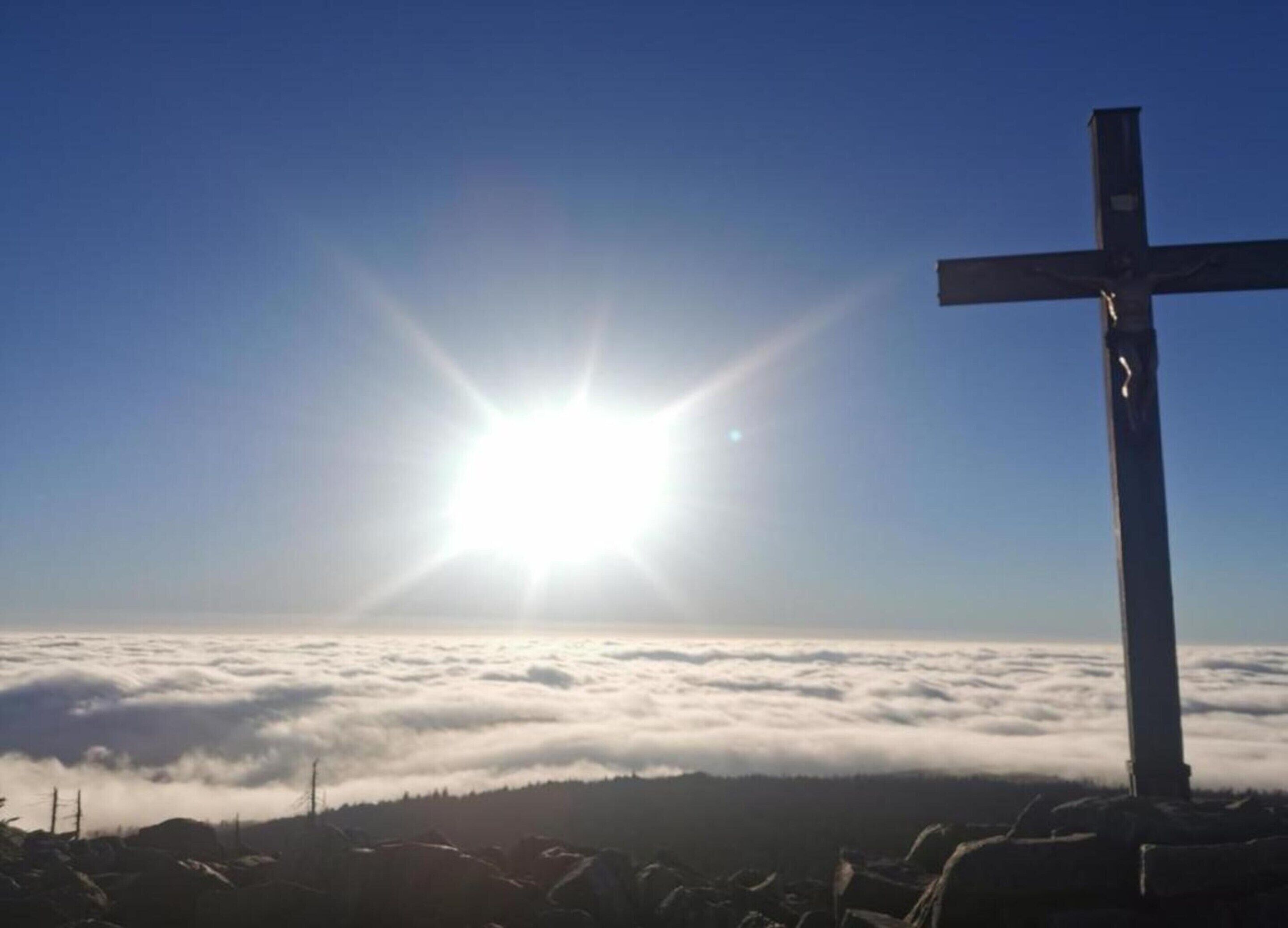 Une croix, avec le soleil brillant derrière les nuages, et un paysage boisé en contrebas.