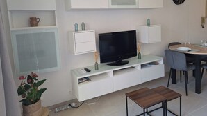 Ein modernes Wohnzimmer mit Flachbildfernseher, weißen Schränken, einem Esstisch mit Stühlen und einer kleinen Topfpflanze.