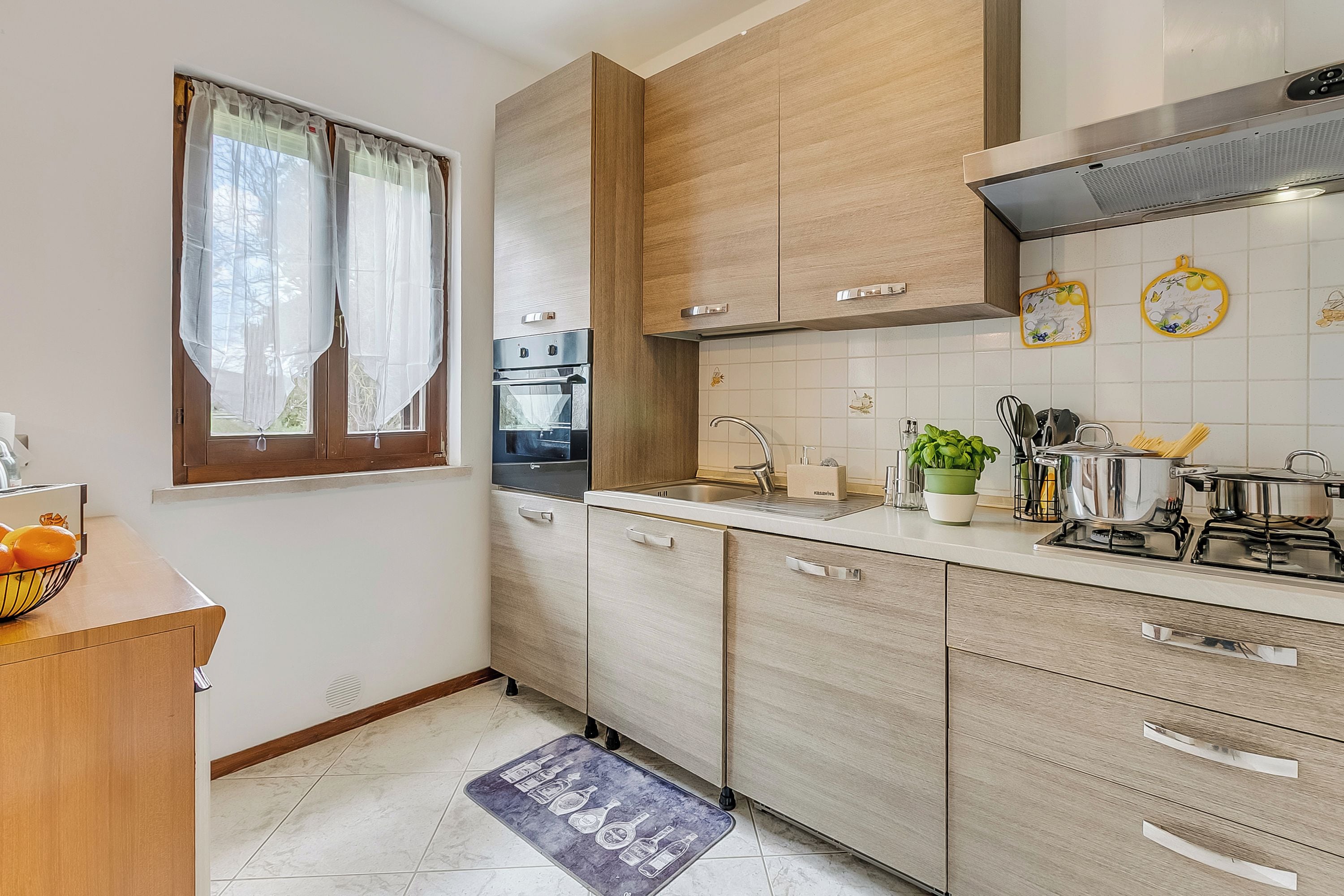 Una cocina moderna con armarios de madera, horno empotrado, cocina a gas y ventana con cortinas ligeras.