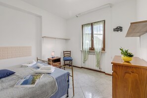 A bedroom with a bed, a chair, a bedside table, a window with curtains, and a small plant on a wooden cabinet. - House Tiziana Close by Mare Argentario (Manciano)