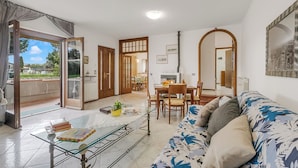 A living room with a sofa, a glass coffee table, a dining area with a table and chairs, and a view through an open door. - House Tiziana Close by Mare Argentario (Manciano)