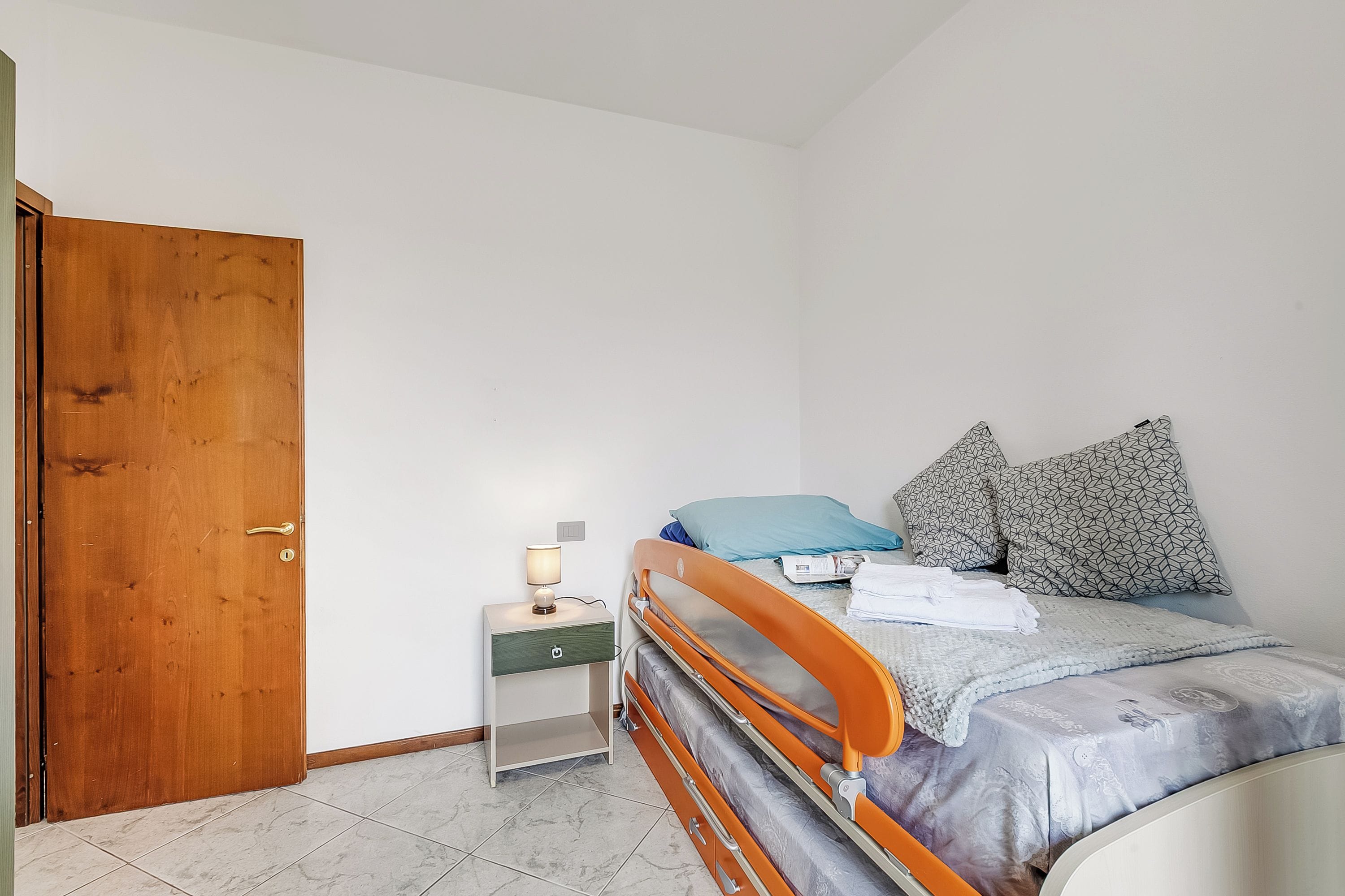 Een slaapkamer met een houten deur, een nachtkastje met een lamp, een bed met kussens en een ladekast.