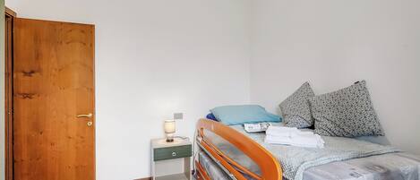Een slaapkamer met een houten deur, een nachtkastje met een lamp, een bed met kussens en een ladekast.