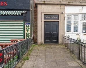 A building with a black door labeled 'Eden View,' a closed shuttered storefront with a sign reading 'Westler HOT DO,' and a metal fence in the foreground. - Eden View Flower Room (Lake District)