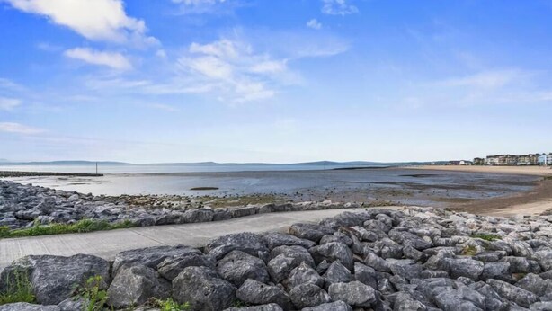 A coastal landscape with a rocky foreground, a concrete path, and a distant shoreline with buildings. - Eden View Jungle Room (Lake District)