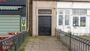 A building with a black door labeled 'Eden View,' a closed shuttered storefront with a sign reading 'Westler HOT DO,' and a metal fence in the foreground. - Eden View Jungle Room (Lake District)