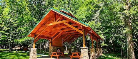 A wooden pavilion with a stone base, surrounded by lush greenery and trees.