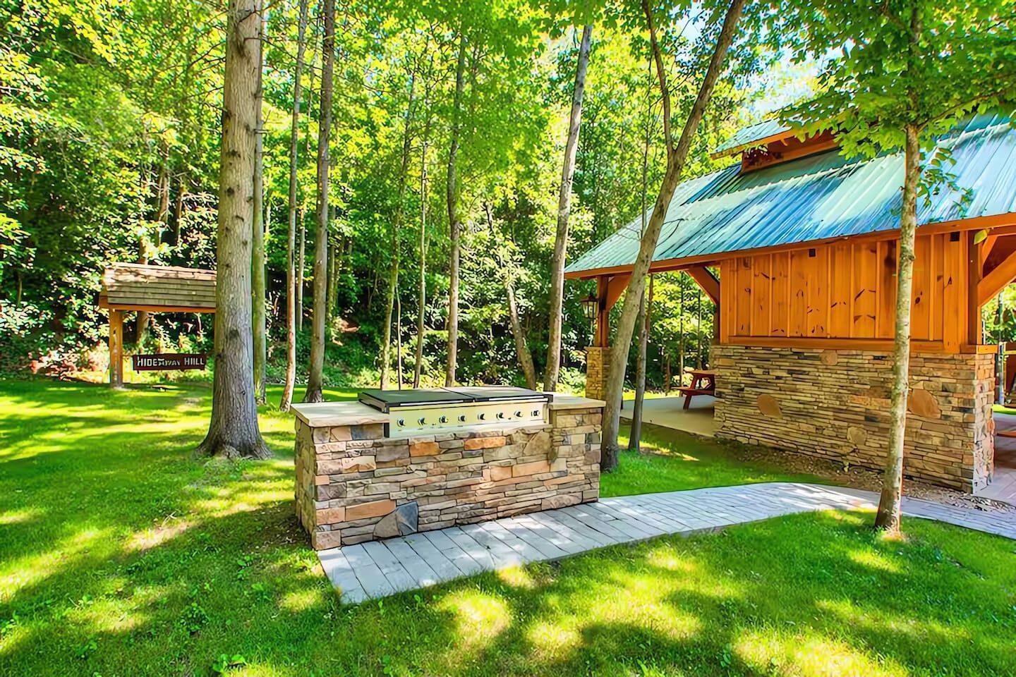 A stone barbecue area with a wooden pergola and a picnic table in a wooded setting.