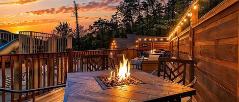 An outdoor deck with a fire pit, wooden furniture, and string lights against a sunset backdrop.