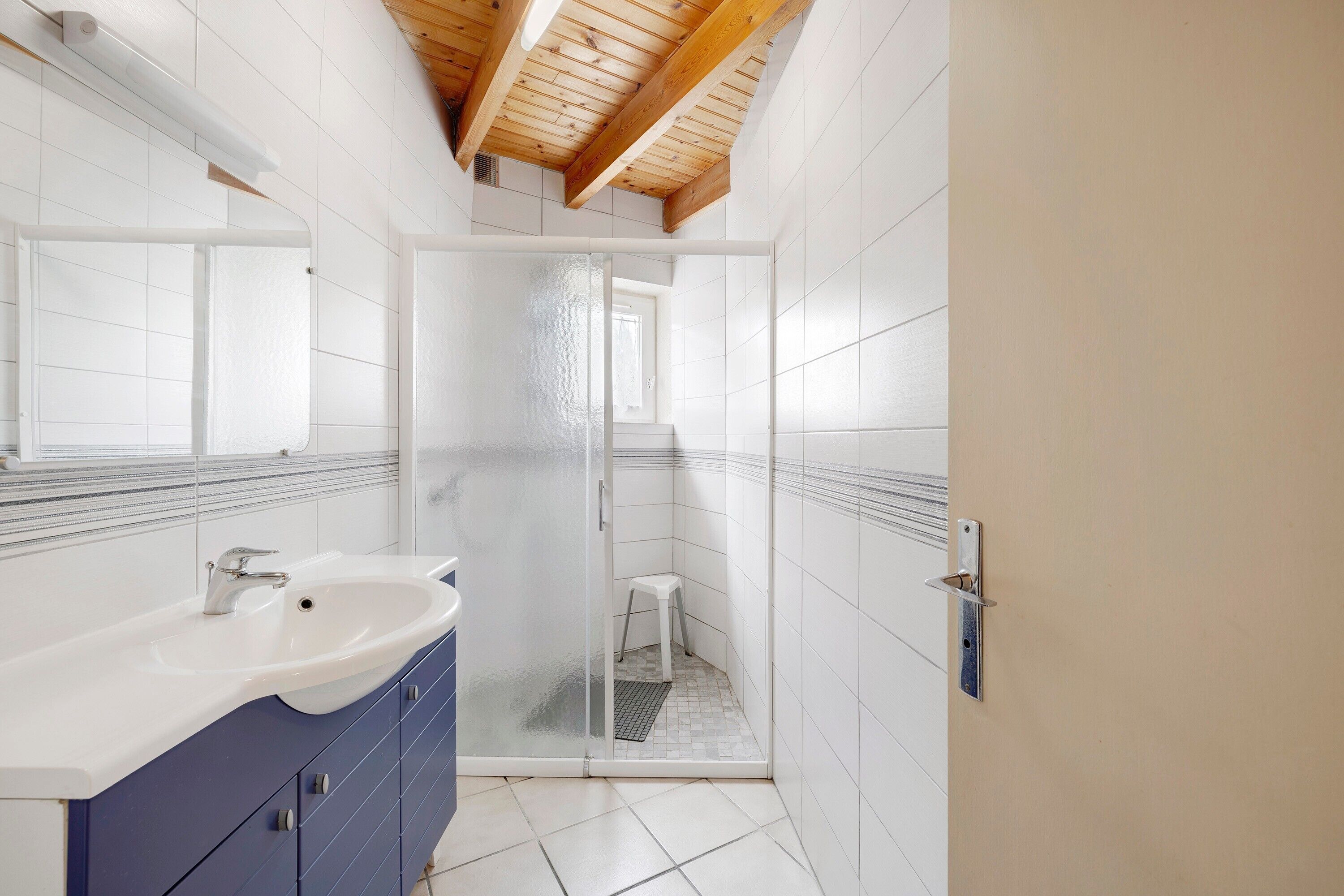Une salle de bain avec un lavabo blanc, des meubles bleus, une douche en verre et un plafond en bois.