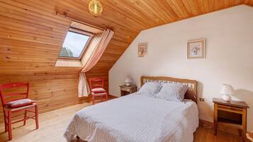 A bedroom with a sloped ceiling, wooden walls, a bed with a patterned bedspread, two nightstands, a window with curtains, and a small picture on the wall.