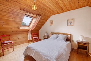 Ein Schlafzimmer mit schrägverdeckter Decke, Holzwänden, einem Bett mit gemustertem Überwurf, zwei Nachttischen, einem Fenster mit Vorhängen und einem kleinen Bild an der Wand.