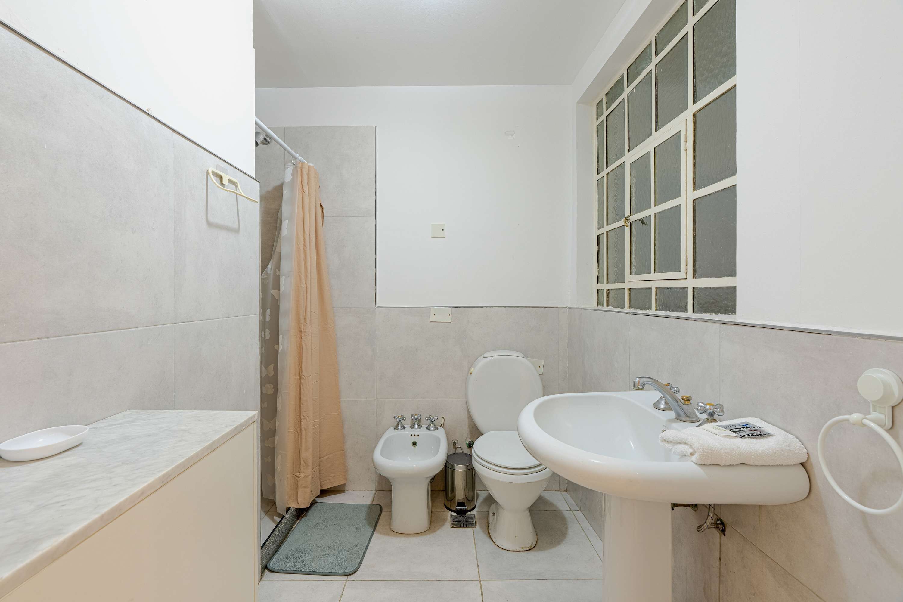 Un baño con inodoro, bidé, lavamanos y una ventana con diseño de celosía.