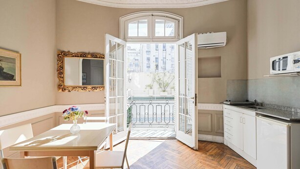 A room with a dining table, chairs, a vase with flowers, a window with open shutters, and a view of buildings outside. - LDV Aparts 2B by MOT Rentals (Buenos Aires)