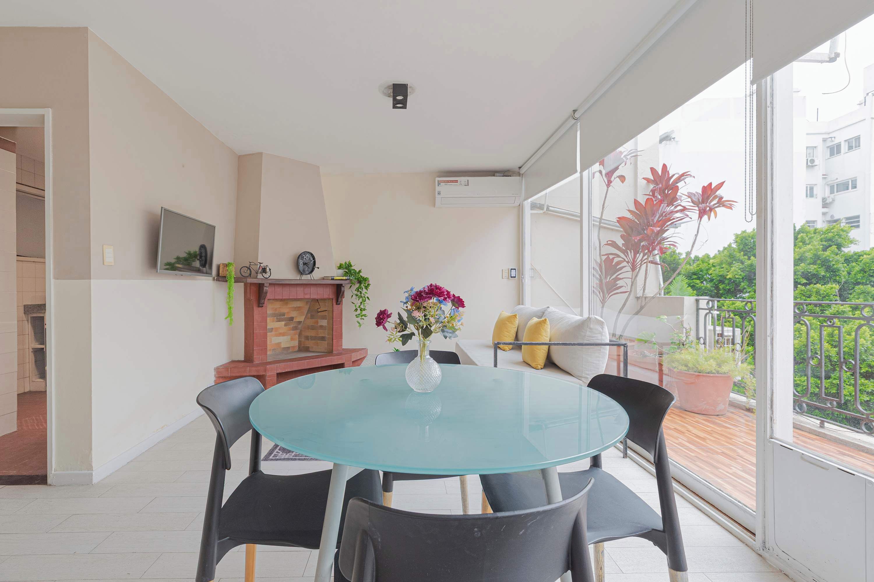 Una sala de estar moderna con una mesa de vidrio, sillas negras, chimenea, televisor y un balcón con plantas en macetas.