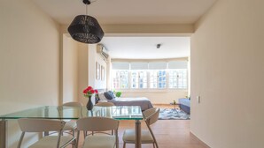 Een eethoek met een glazen tafel en stoelen, een zwart hanglamp en uitzicht op een slaapkamer met een bed en raam.