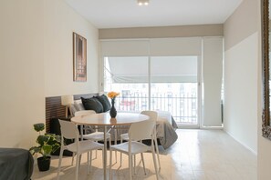 A modern living room with a round dining table, white chairs, a sofa, a potted plant, and a large window with a view. - LDV Aparts 5B by MOT Rentals (Buenos Aires)