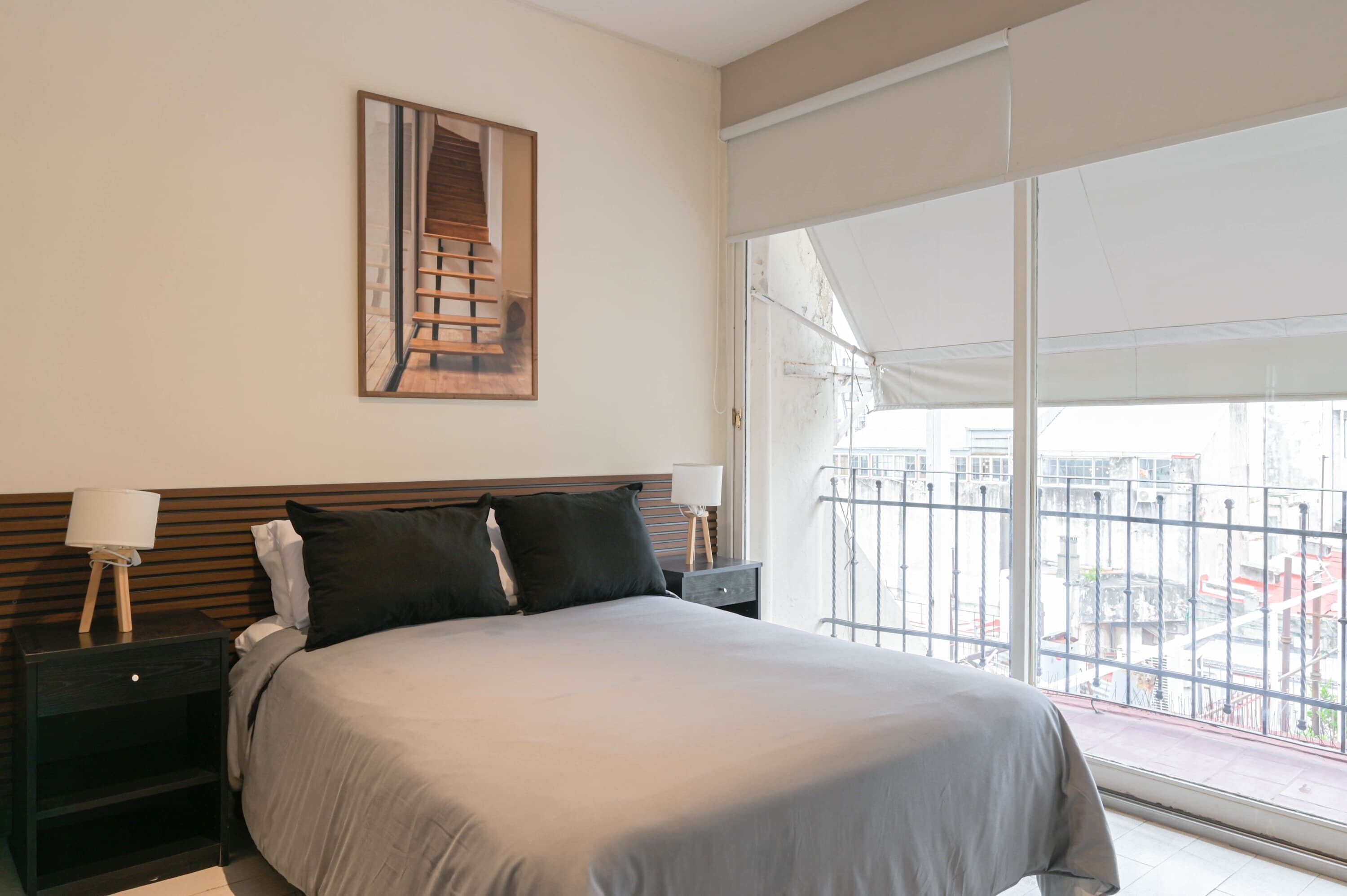 Un dormitorio con cama, mesita de noche, lámpara, cuadro en la pared y un amplio ventanal con vistas a edificios.
