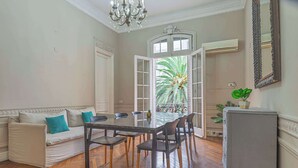 A dining area with a glass table, chairs, a sofa with cushions, a chandelier, and a view of a palm tree through a window. - LDV Aparts 2A (2 Bedrooms) by MOT Rentals (Buenos Aires)