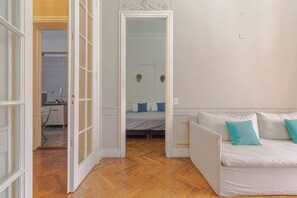 Ein Zimmer mit Holzboden, einem weißen Sofa mit türkisfarbenen Kissen und einem Bett mit blau-weißen Kissen.