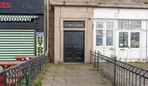 A building with a black door labeled 'Eden View,' a closed shuttered storefront with a sign reading 'Westler HOT DO,' and a metal fence in the foreground. - Eden View Jasmine Room (Lake District)
