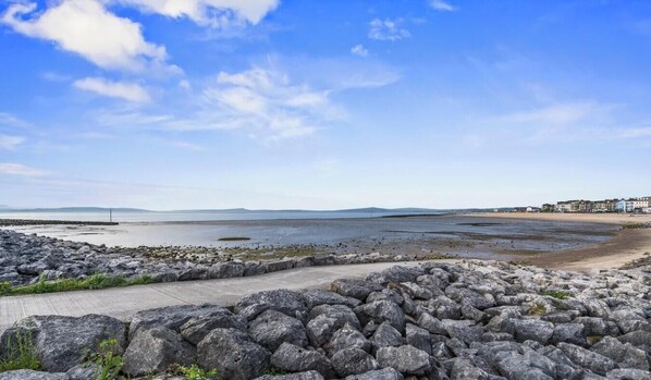A coastal landscape with a rocky foreground, a concrete path, and a distant shoreline with buildings. - Eden View Jasmine Room (Lake District)