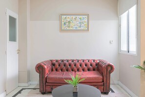 A red leather sofa, a framed abstract painting, and a potted plant on a round table in a room with a door and a window. - LDV Aparts 2F by MOT Rentals (Buenos Aires)