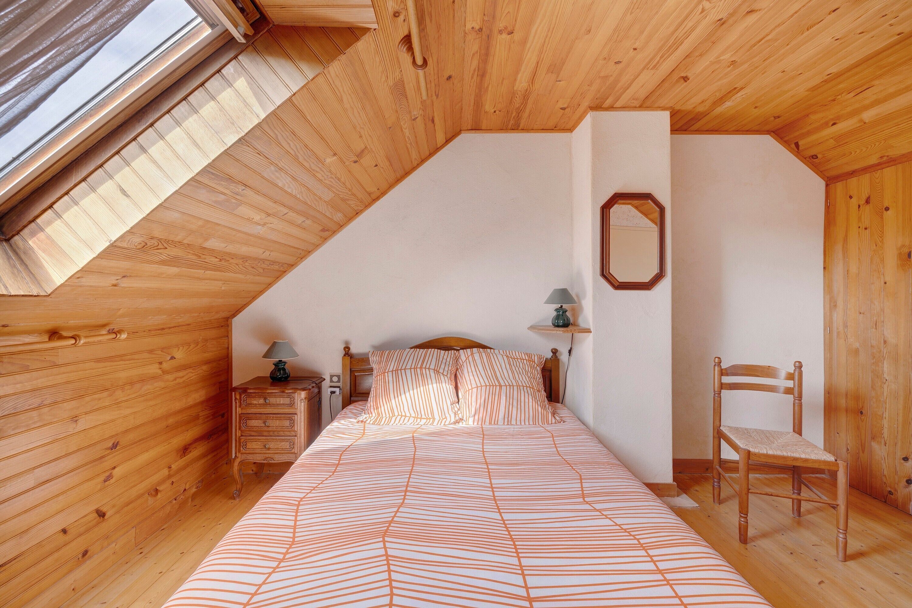 A bedroom with a sloped ceiling, wooden walls, a bed with striped bedding, a nightstand, a chair, and a mirror on the wall.