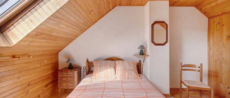 A bedroom with a sloped ceiling, wooden walls, a bed with striped bedding, a nightstand, a chair, and a mirror on the wall.