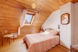 Een slaapkamer met een schuine kap, dakraam, een bed met gestreepte beddengoed, een stoel, een nachtkastje met een lamp, en een spiegel aan de muur.