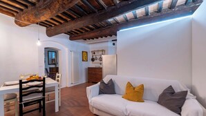 Ein Wohnzimmer mit einem weißen Sofa, einem Esstisch mit Stühlen, einer Küchenzeile mit Kühlschrank und einer rustikalen Decke.