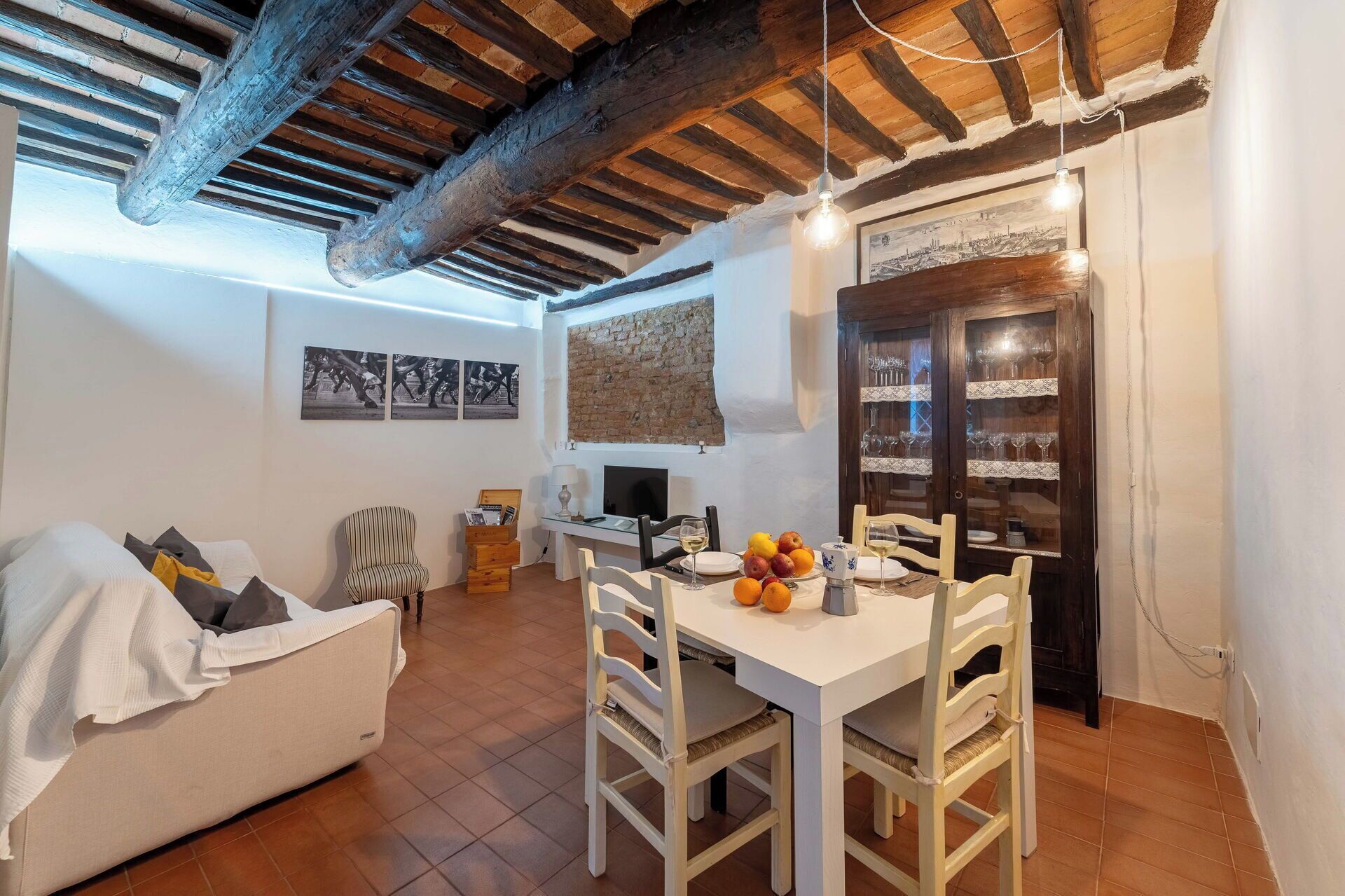 A cozy living room with a dining area, wooden beams on the ceiling, a TV, and a brick wall.
